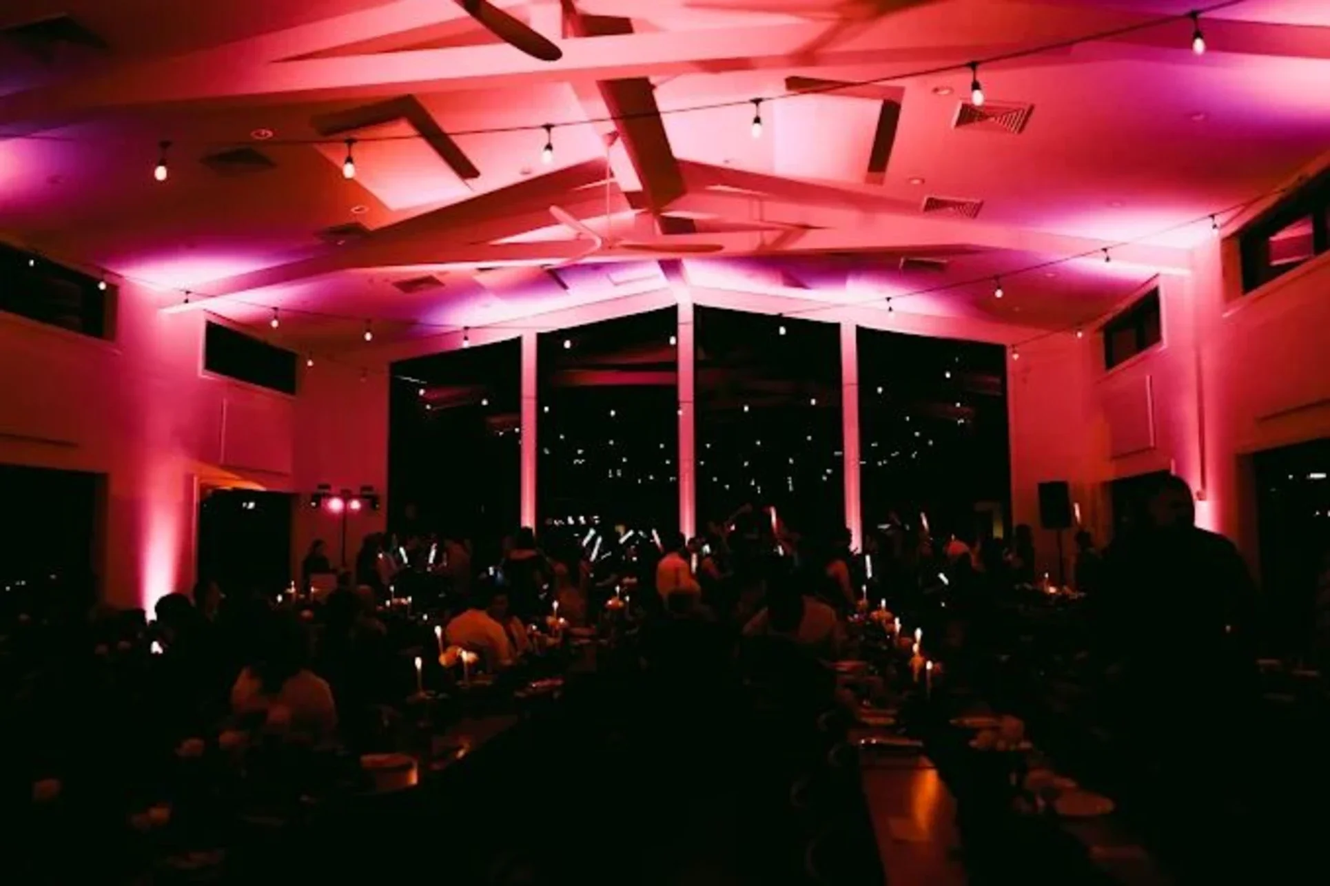 Wedding reception bathed in pink uplighting with guests holding LED party sticks under string lights
